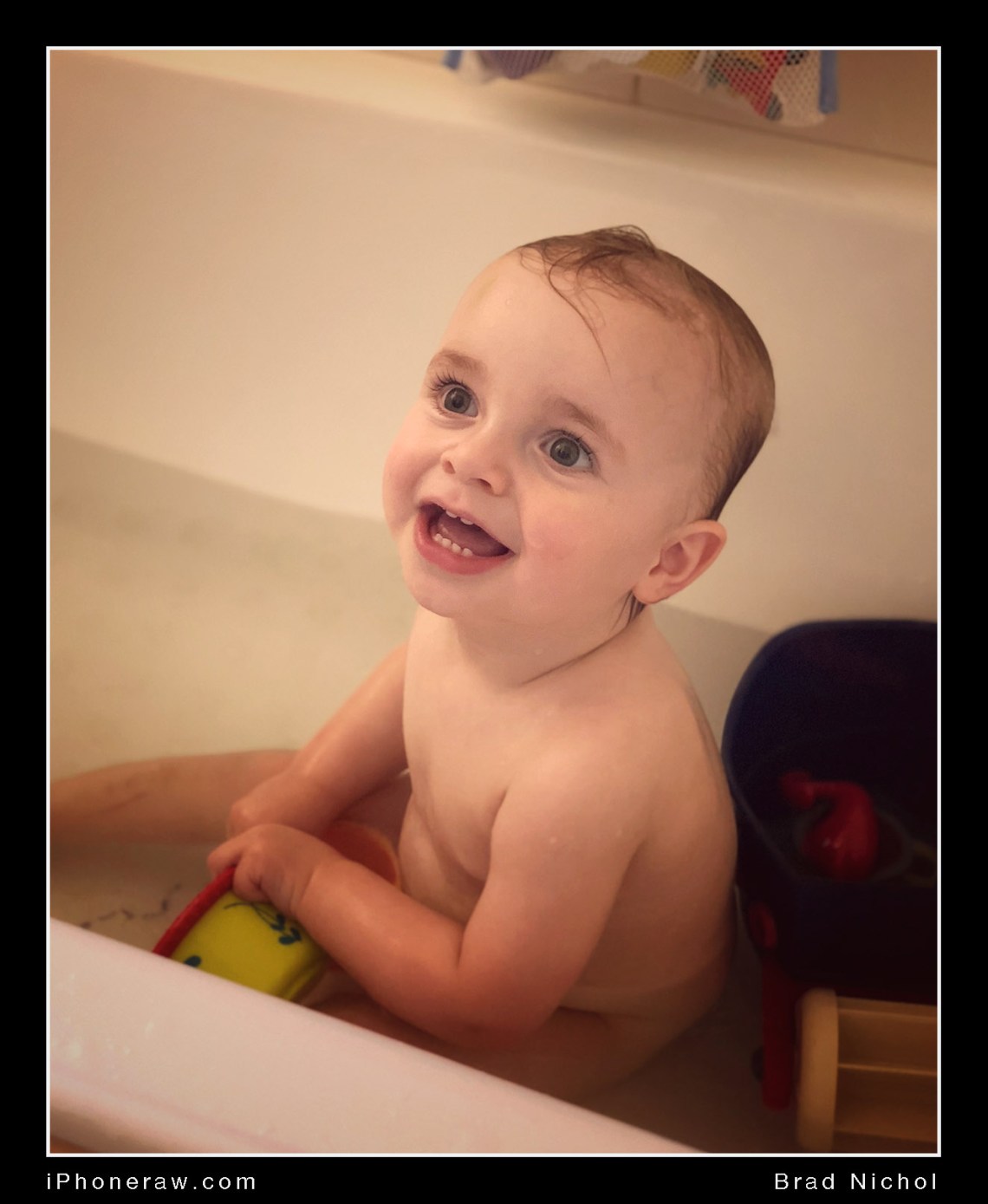 Baby in bath with toys, looking up and smiling, sepia low saturation colour tint, taken iPhone X portrait mode.